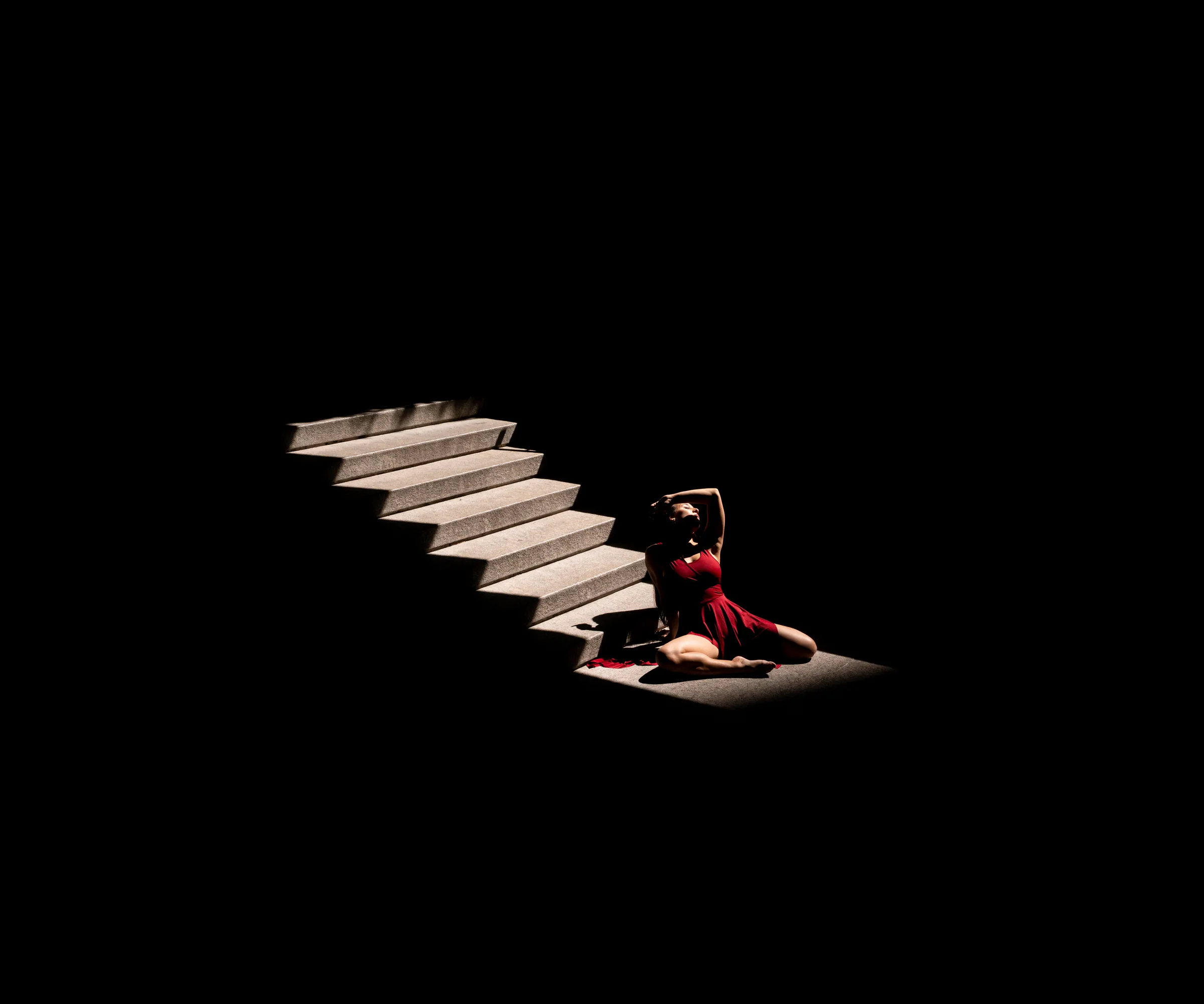 Bailarina recostada bajo una escalera iluminada en un espacio oscuro